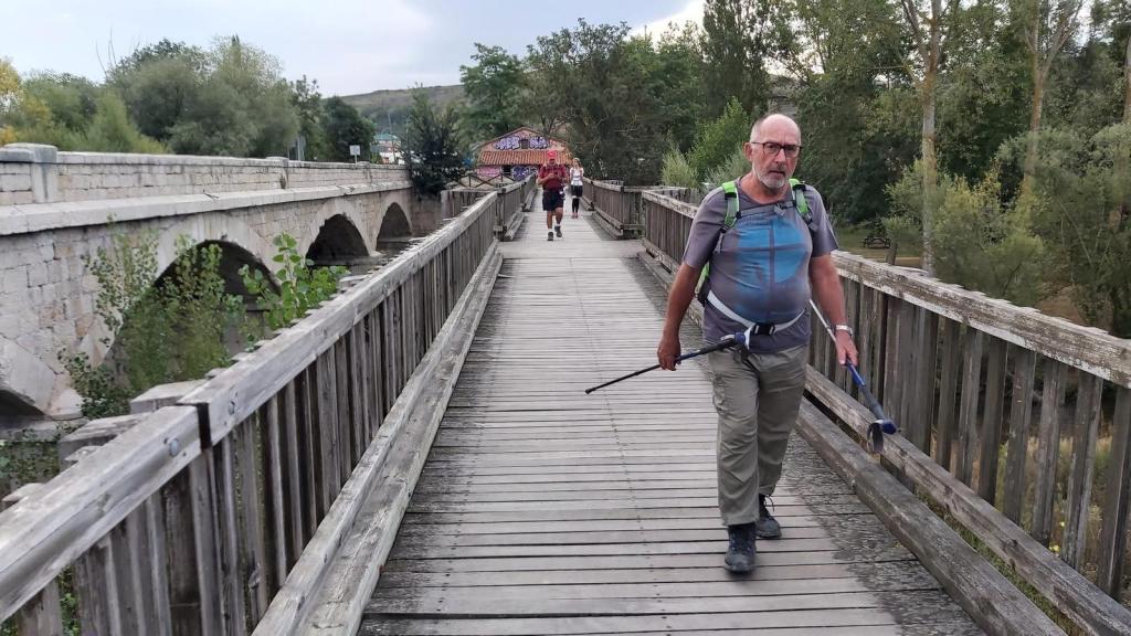 Un peregrino cruza un puente de madera mientras realiza la ruta 'Etapa 11: Belorado-Agés' del Camino de Santiago, con árboles y vegetación en el fondo.