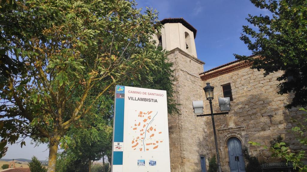 Señal de dirección del Camino de Santiago en Villambistia, con un paisaje de árboles y un edificio histórico al fondo.