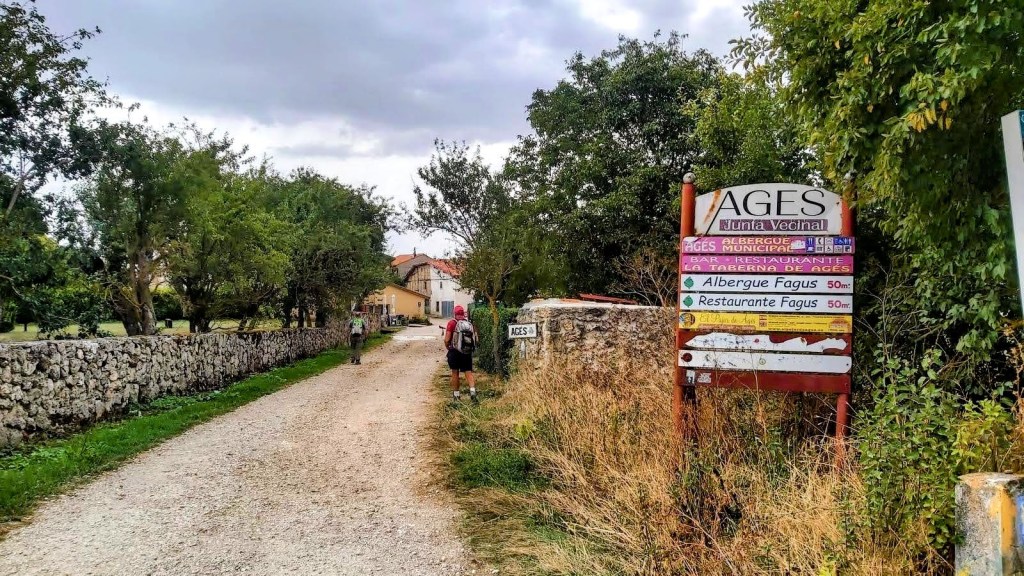 Entrada a Agés con un cartel que señala dirección a Albergue y Restaurante Fagus, rodeada de vegetación y un camino de tierra.