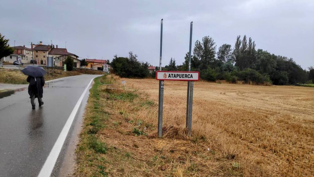 Señal de bienvenida a Atapuerca en un día lluvioso, con un caminante bajo un paraguas en un camino junto a campos de trigo.