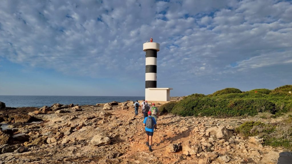 S'ESTANYOL a VALLGORNERA (Cala Pi) - Caminando por Mallorca Grupo de caminantes acercándose al Faro de s'Estalella, rodeado de rocas y vegetación, con un cielo nublado sobre el mar.