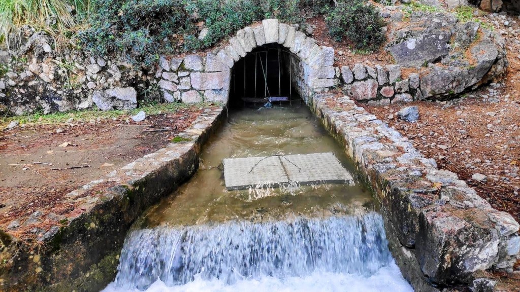 Font des Verger con agua fluyendo y estructuras de piedra, rodeada de vegetación en un entorno natural.