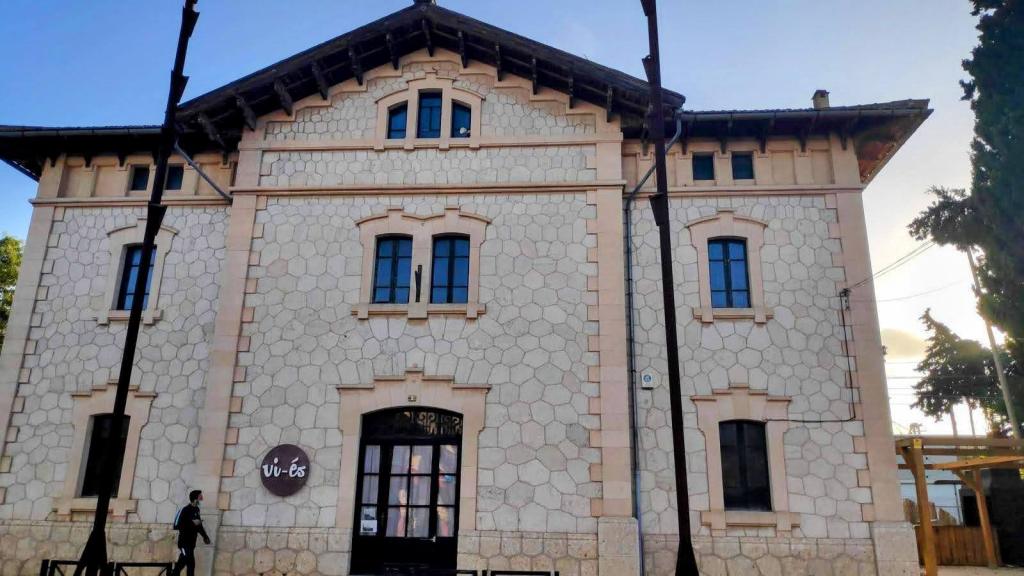 Edificio de la estación del tren de Sineu, de estilo arquitectónico notable con fachada de piedra clara, ventanas simétricas y detalles decorativos en la parte superior, situado en un entorno urbano.