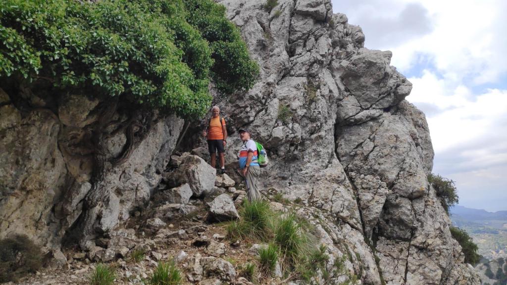Dos excursionistas avanzan por el Pas de Sa Partió o Pas de s'Heura, rodeados de vegetación y peñascos en un entorno montañoso.