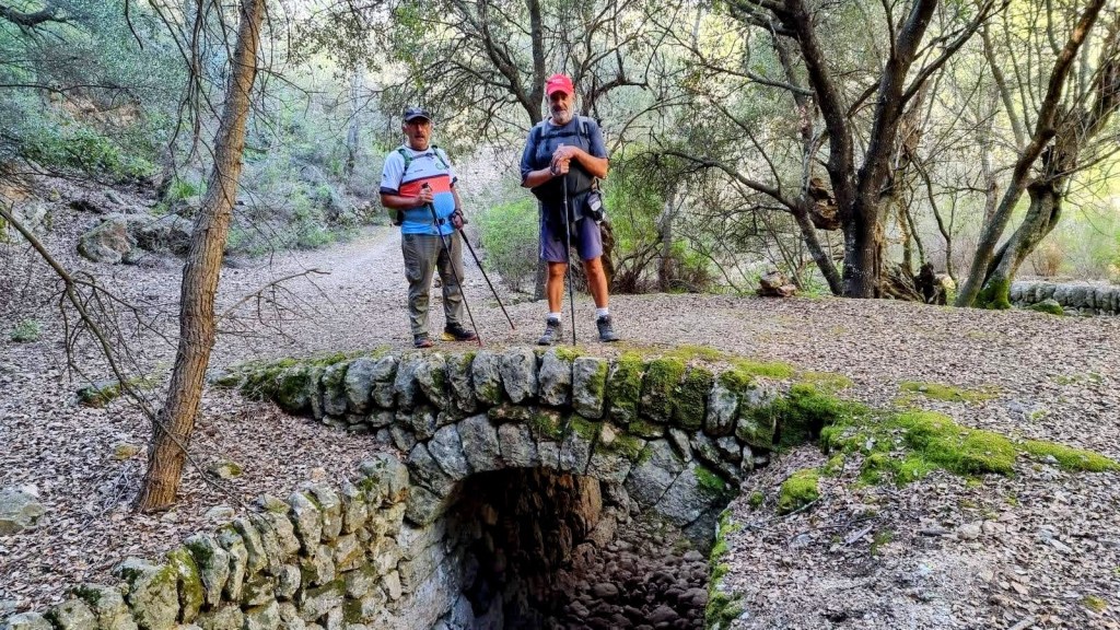 Dos excursionistas de pie sobre un puente de piedra en un sendero forestal, rodeados de árboles y vegetación.