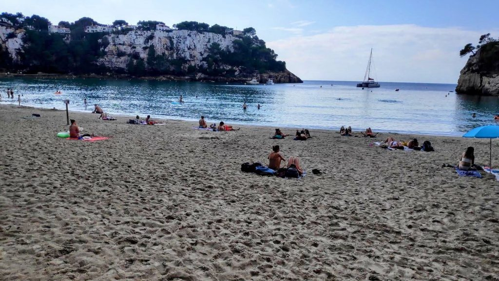Playa de Cala Galdana con turistas relajándose en la arena, fondo de acantilados y un barco navegando en el agua.