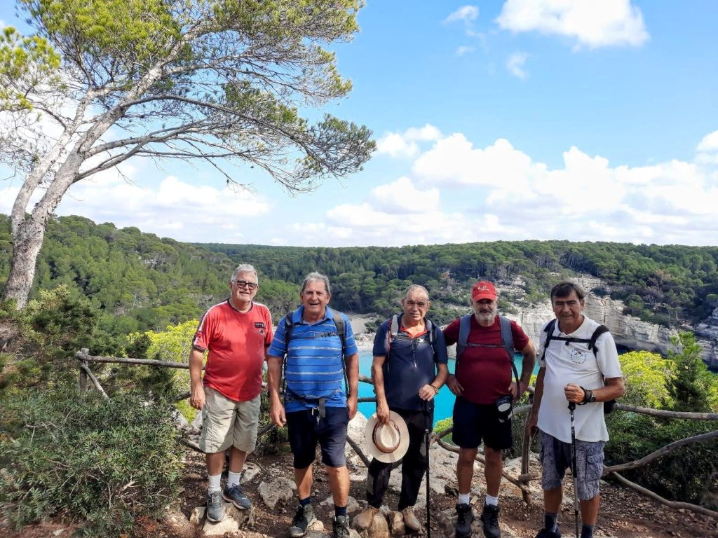 Grupo de cinco senderistas en un mirador, frente a un paisaje de vegetación y un barranco con vistas al mar, en la ruta Camí de Cavalls en Menorca.
