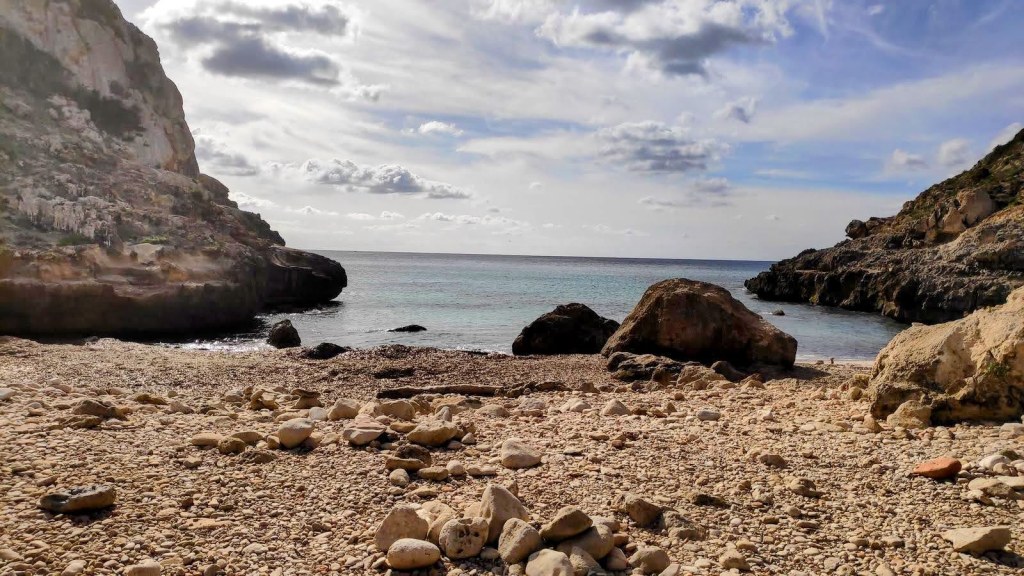 Vista de una playa rocosa con grandes piedras y un mar tranquilo en el horizonte, rodeada de acantilados y una atmósfera serena.