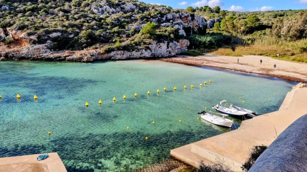 Vista panorámica de Cala Sant Esteve con aguas claras y tranquilas, rodeada de vegetación y rocas, y con dos barcos atracados en la orilla.