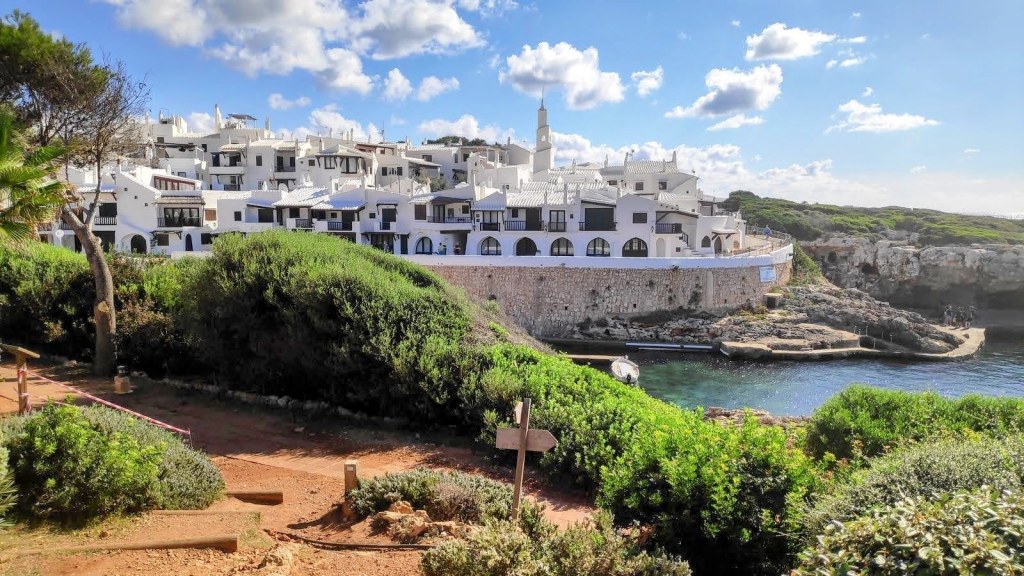 Vista de Binibequer, con casas blancas y vegetación en primer plano, junto a una pequeña cala.