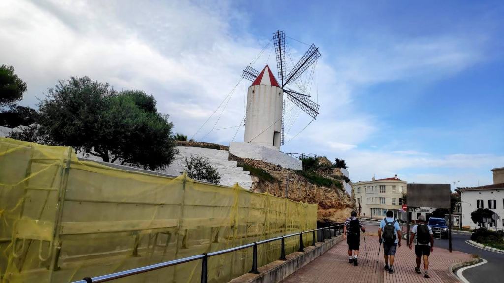 Con tres personas caminando por un camino junto a un molino de viento con aspas grandes y un edificio al fondo, en un día parcialmente nublado.
