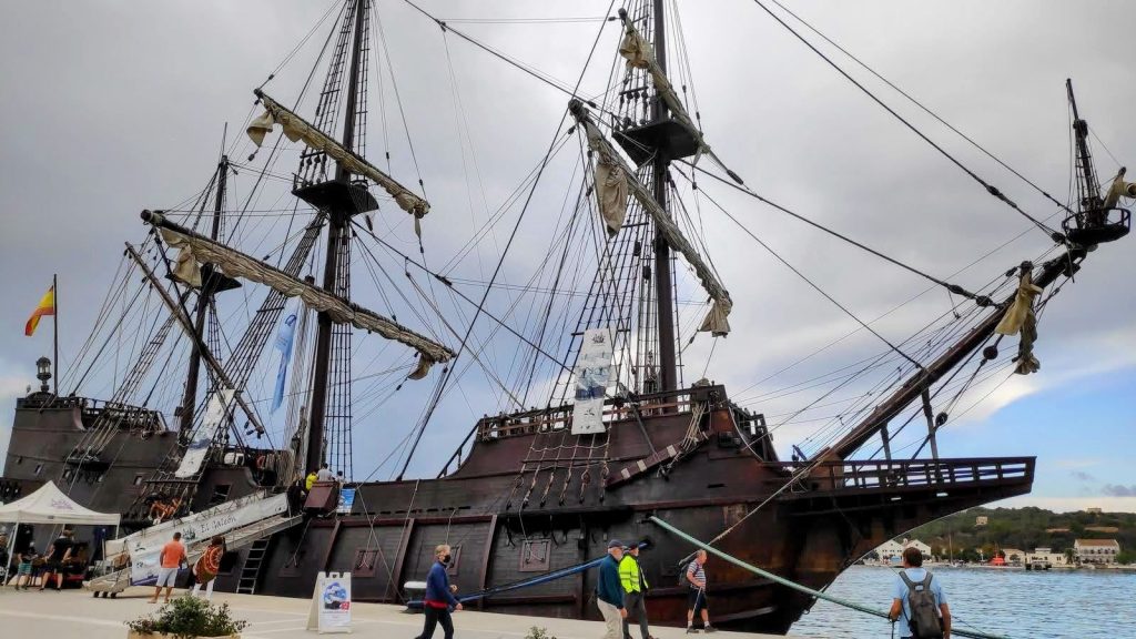 Barco antiguo atracado en el Puerto de Mahón, con velas y mástiles visibles, rodeado de personas que caminan cerca.