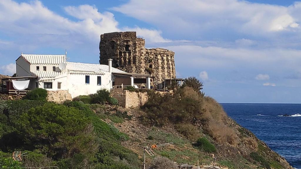 Vista de la Torre de Sa Mesquida en un acantilado junto al mar, con una casa blanca en primer plano y un cielo parcialmente nublado.