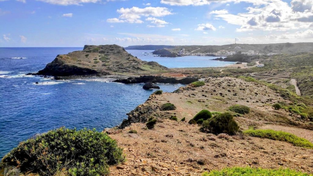 Panorámica de un paisaje costero que muestra acantilados, vegetación baja y un mar azul en calma, con pequeños pueblos visibles en el fondo.