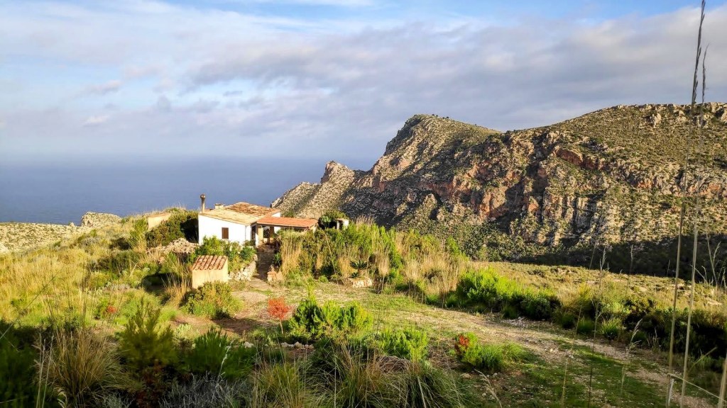 Vista de una casa rural rodeada de vegetación en la costa oeste de Mallorca, con montañas y el mar al fondo bajo un cielo nublado.