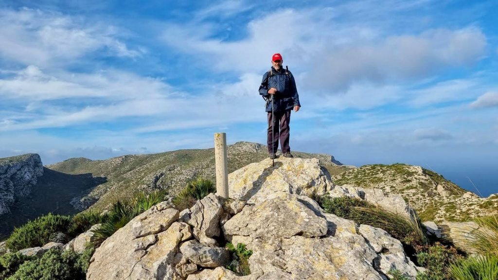 Hombre de pie sobre una roca en la cima del Puig de Macià Andreu, con un hito geodésico, rodeado de un paisaje montañoso y un cielo parcialmente nublado.