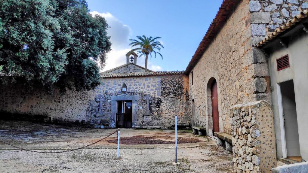 Vista de la entrada de la Ermita de la Santísima Trinidad, con paredes de piedra y un cielo nublado detrás. Se observan árboles y una palmera cerca de la entrada.