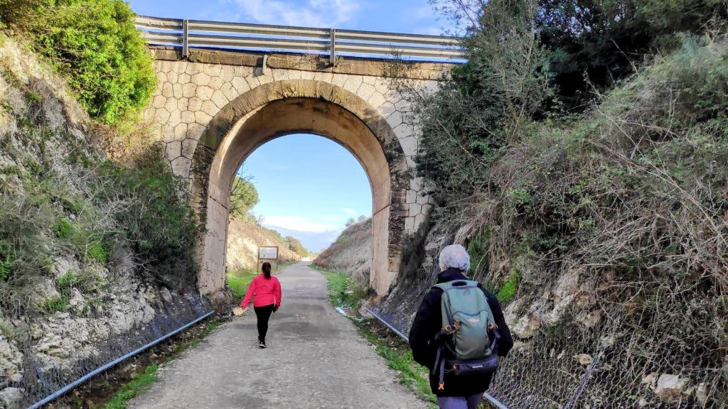 Personas caminando por un camino de tierra bajo un puente arqueado, vegetación a los lados en un día despejado.