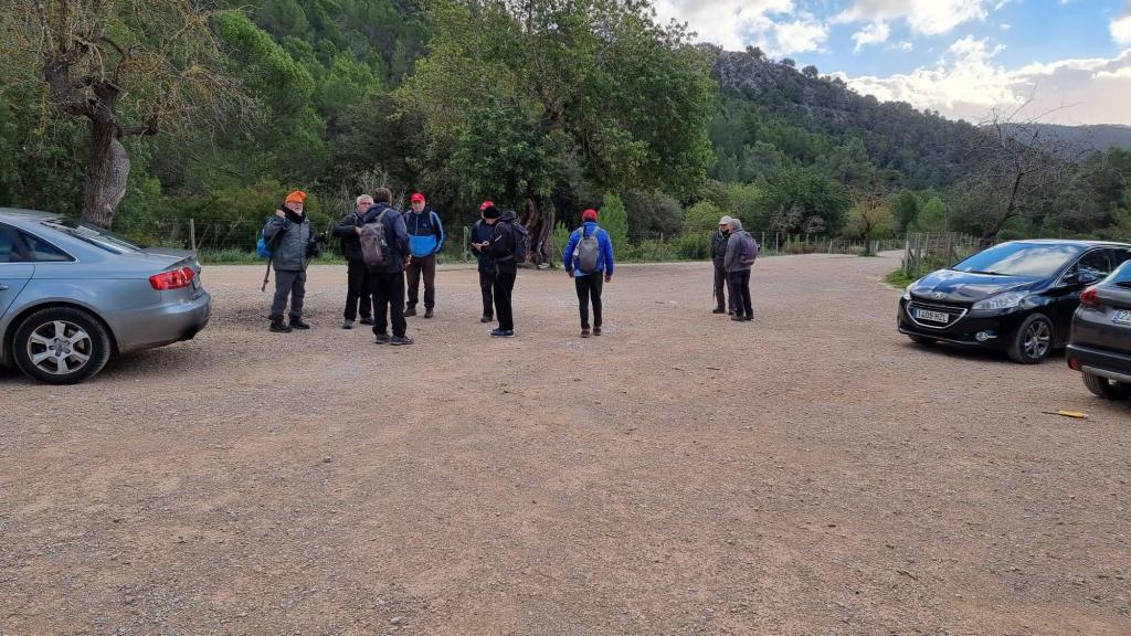 Grupo de personas reunidas en un aparcamiento rural, rodeados de árboles y montañas, preparándose para iniciar una ruta de senderismo.