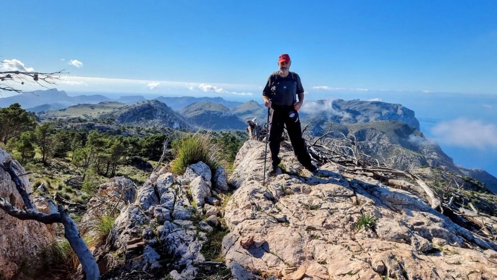 Hombre de pie en la cima de una montaña, rodeado de vistas panorámicas de la sierra, árboles y rocas.