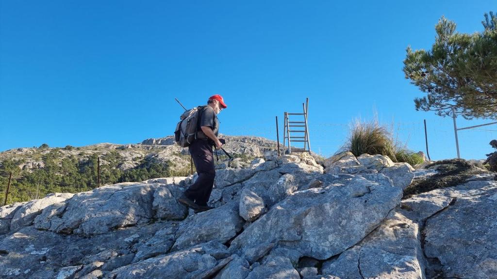 Una persona camina sobre un terreno rocoso mientras asciende, con una escalera visible y un paisaje de montañas al fondo bajo un cielo azul claro.