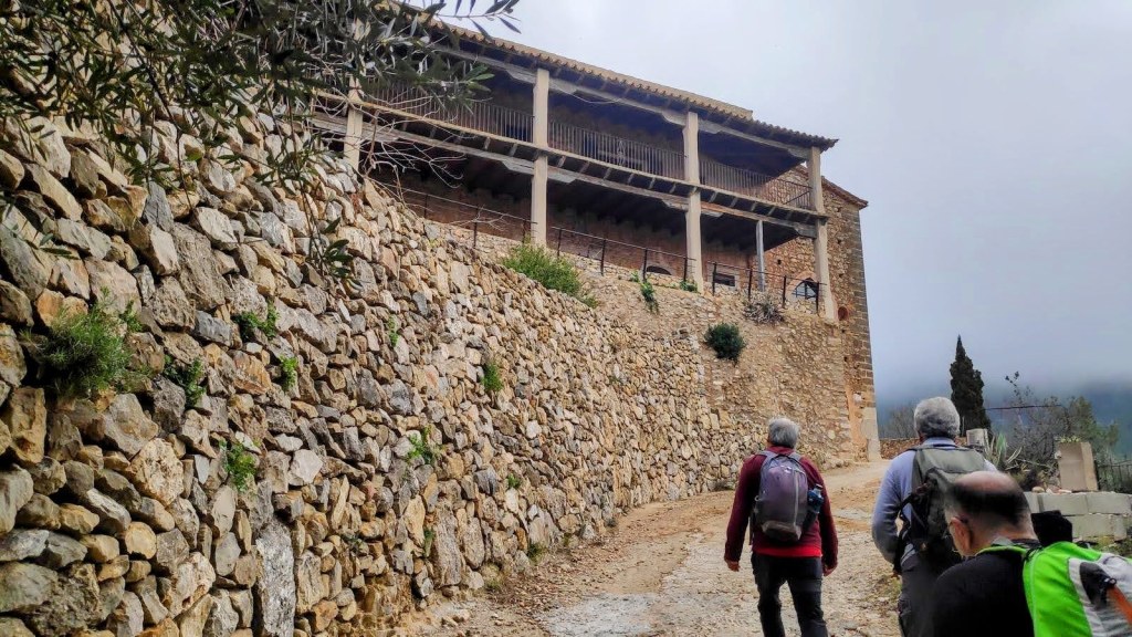 Grupo de senderistas caminando por un camino de piedra hacia un edificio de dos pisos con un balcón, rodeado de un paisaje montañoso y vegetación.