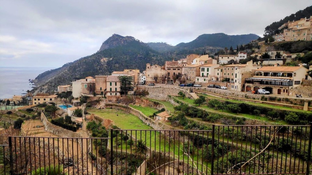 Vista panorámica de Banyalbufar, con casas y edificios en la ladera de una montaña, colinas verdes y el mar al fondo, bajo un cielo nublado.