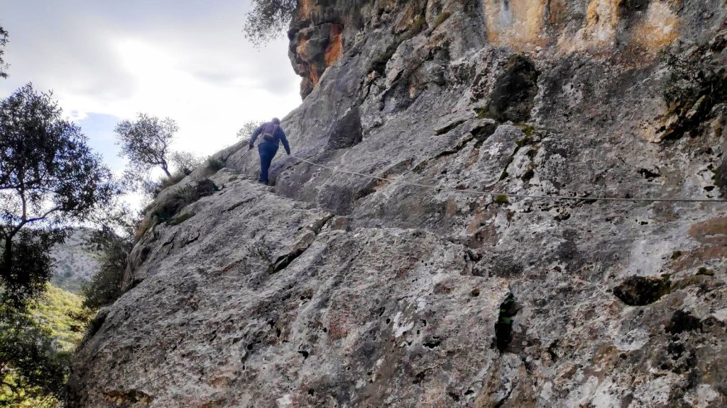 Un senderista ascendiendo por una roca utilizando una cuerda de seguridad, rodeado de vegetación en un paisaje montañoso.