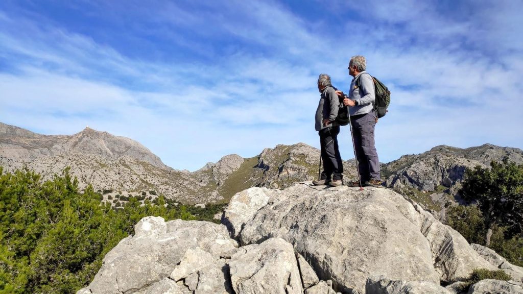 SERRA d'AMOS desde Son Ordinas - Caminando por Mallorca Dos senderistas contemplando el paisaje montañoso desde una roca en la Serra d'Amos, Mallorca, bajo un cielo azul con nubes dispersas.
