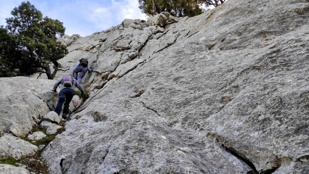 SERRA d'AMOS desde Son Ordinas - Caminando por Mallorca Dos personas subiendo por una pendiente rocosa en la Serra d'Amos, con un fondo de cielo azul y vegetación.