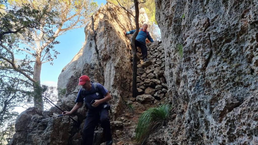 Dos personas escalando por un terreno rocoso en un sendero montañoso, rodeados de árboles y vegetación.