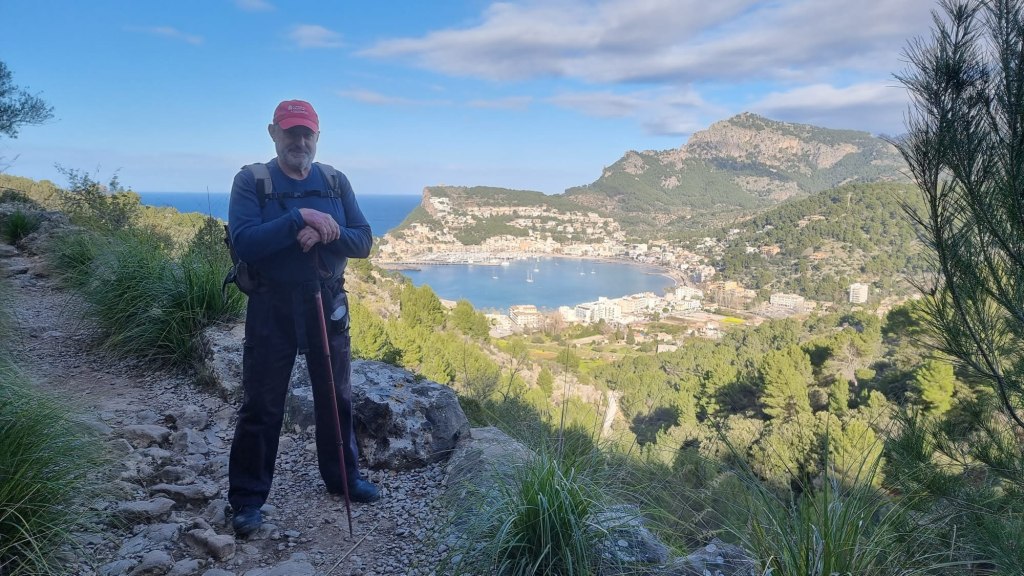 Hombre con bastón parado en un sendero montañoso con vistas al puerto de Sóller y las montañas circundantes.