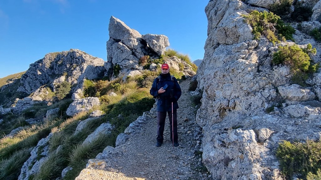 Hombre de pie en un sendero rocoso rodeado de grandes rocas en un paisaje montañoso, posiblemente en la Ruta Es Cairats-Pla dels Aritges.
