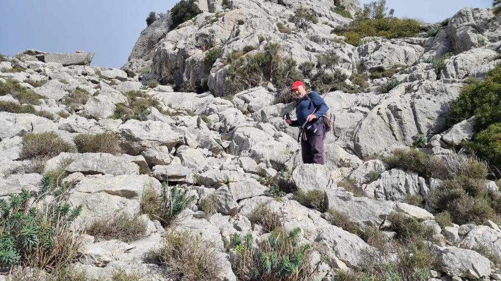 Hombre caminando entre rocas en un terreno accidentado de la Serra des Cairats, con vegetación en el fondo.