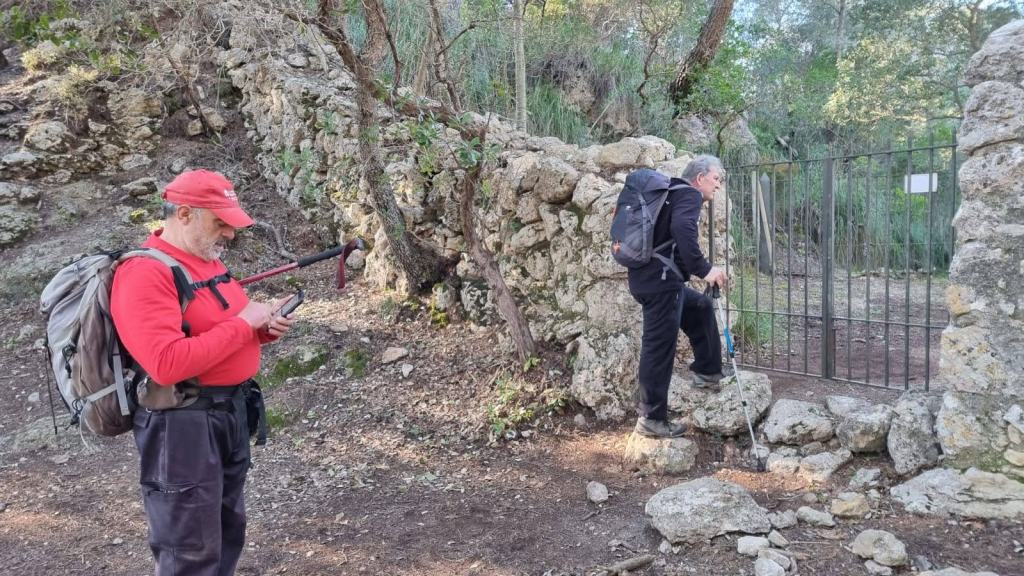 Dos excursionistas en un sendero en el bosque, uno revisando su teléfono móvil y el otro subiendo por unas rocas junto a una puerta de metal.