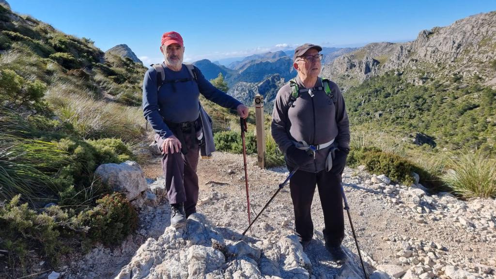 Dos senderistas en el Coll des Prat, sobre un camino rocoso con paisajes de montañas verdes y cielos despejados de fondo.