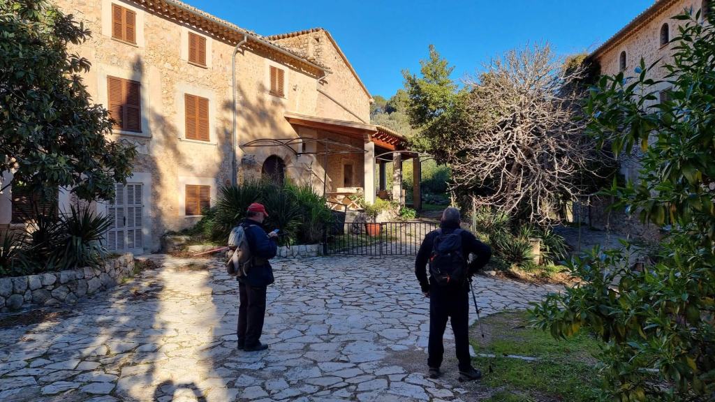 Dos caminantes en un camino empedrado frente a Can Jeroni Gros y Can Ques en la ruta Camí de Rocafort-Camí de Castelló-Camí de Son Sales, rodeados de vegetación.