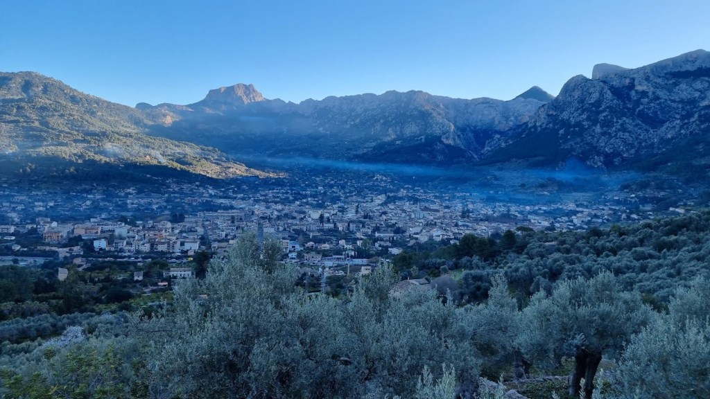 Vista panorámica de Sóller, Mallorca, con montañas y olivares en primer plano, bajo un cielo despejado.