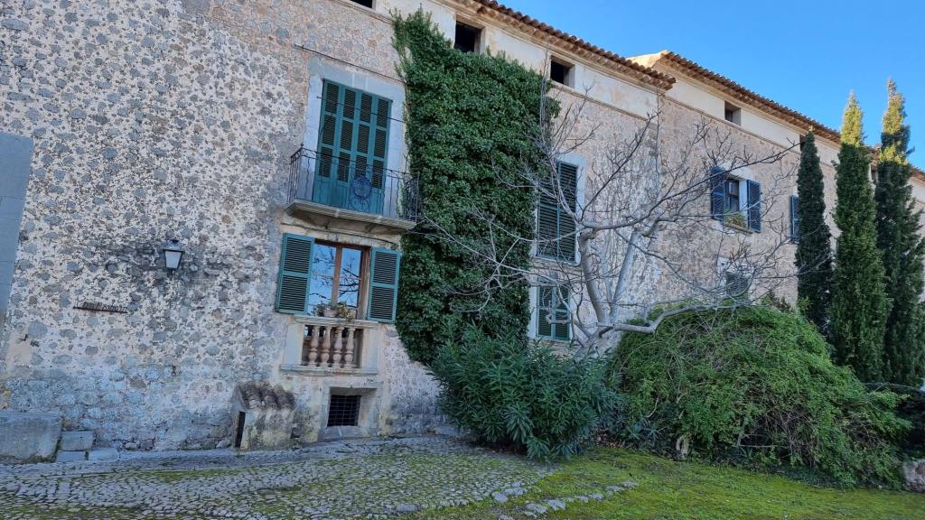 Fachada de una antigua casa de piedra con ventanas de persianas verdes rodeada de vegetación en un claro día soleado.