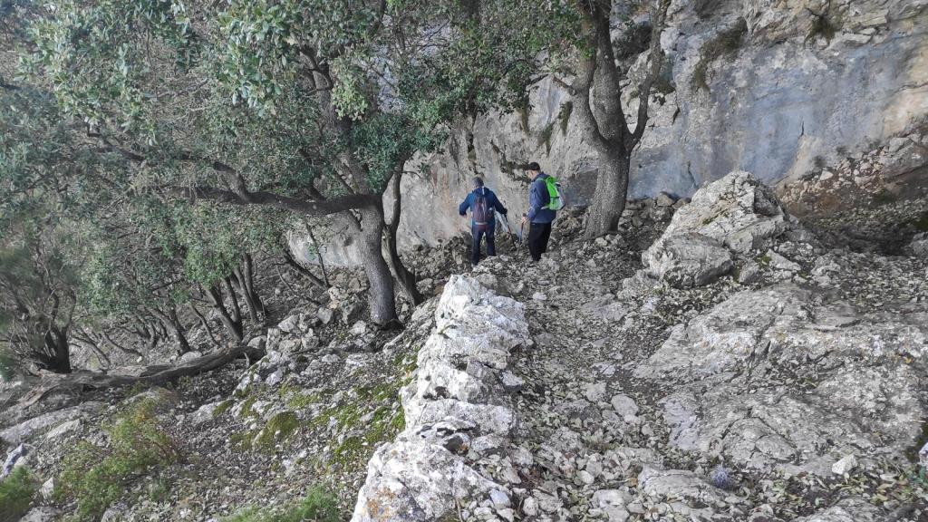 Dos excursionistas caminan por un sendero rocoso en los cingles o riscos de Son Rul-lan , rodeados de árboles y rocas, durante la Ruta de la Pedra en Sec-GR221-Etapa 5.