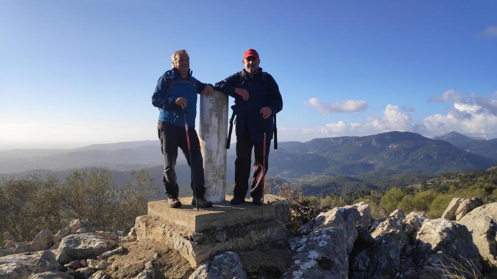 Dos personas posando en la cima del Puig des Boixos, con un vértice geodésico en el centro, rodeados de un paisaje montañoso bajo un cielo despejado.