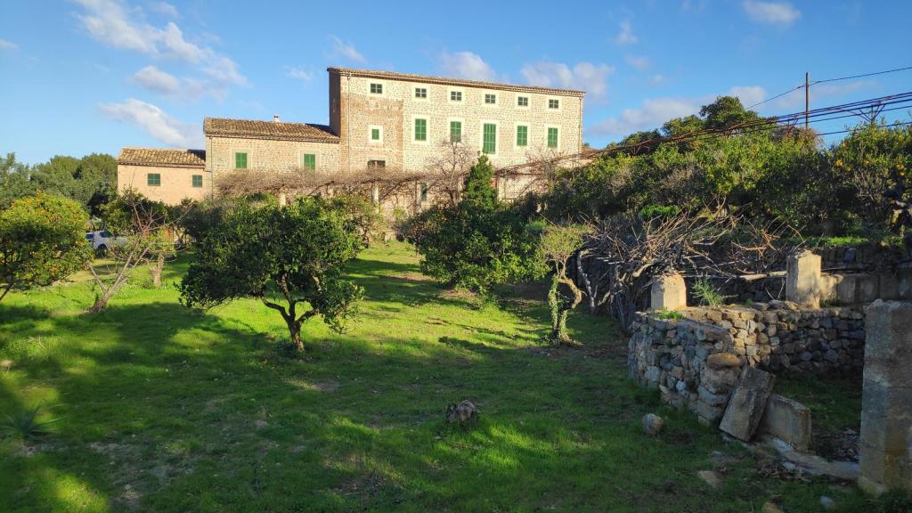 Vista de una antigua finca agrícola con paredes de piedra y ventanas verdes, rodeada de árboles frutales y un amplio jardín de césped bajo un cielo parcialmente nublado.