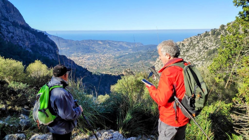 Ruta de la Pedra en Sec-GR221-Etapa 7 - Caminando por Mallorca Dos excursionistas observando el paisaje desde un sendero en la 'Ruta de la Pedra en Sec-GR221-Etapa 7', con vistas al valle de Sóller y el mar al fondo.