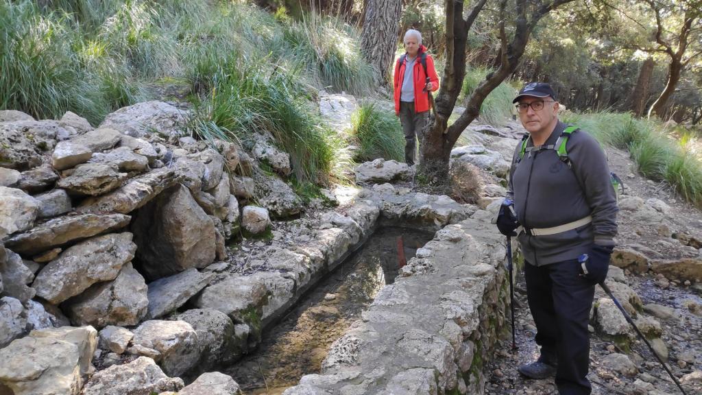 Personas disfrutando de un paisaje natural cerca de la font d'en Gallina en la Ruta de la Pedra en Sec-GR221-Etapa 8, con vegetación densa y rocas en el entorno.
