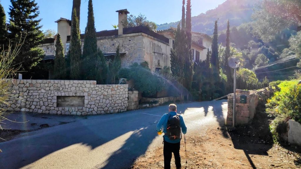 Hombre caminando por una carretera rural, con una casa de piedra y cipreses en el fondo, en un paisaje de montañas.