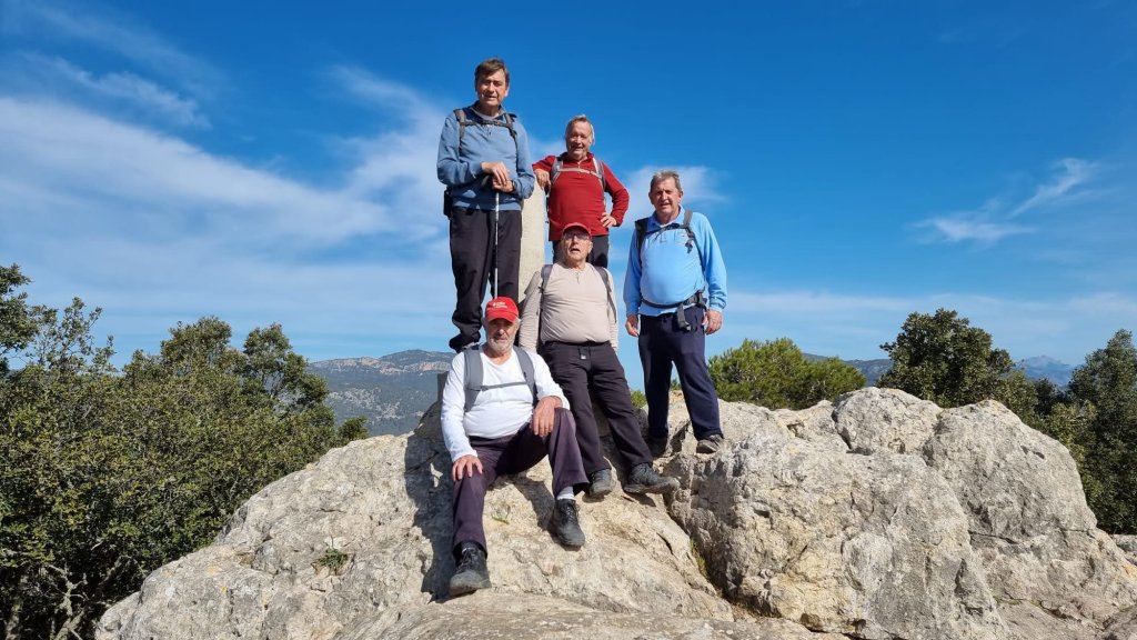 Grupo de cinco personas de pie y sentadas sobre una roca en la cima del Puig de Na Bauçana, con un paisaje montañoso al fondo y un cielo azul despejado.