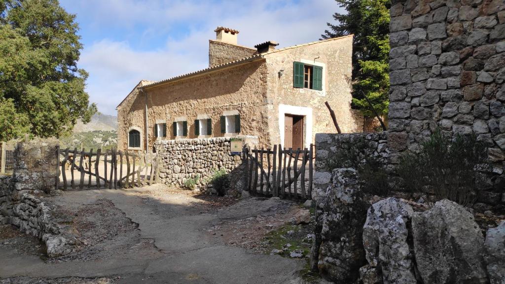 Vista de las Casas de Binifaldó, un edificio de piedra con ventanas verdes, rodeado de naturaleza en un día nublado, con una puerta de entrada de madera y un camino que lleva a la estructura.