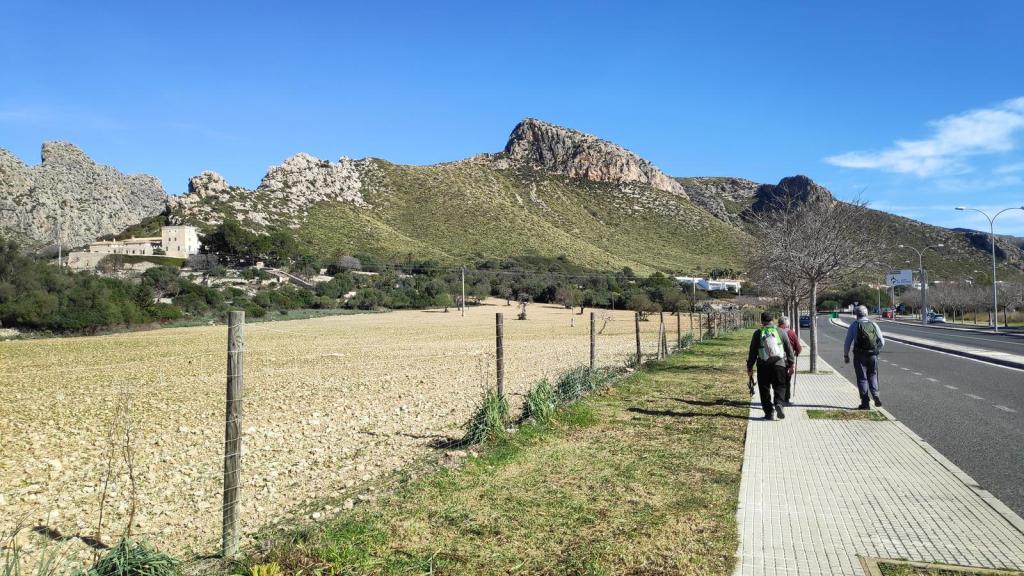 Camino pavimentado junto a un campo de cultivo y montañas en el fondo, con tres personas caminando por el sendero.