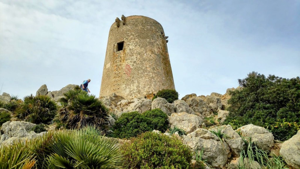 Vista de la Talaia d'Albercutx, una antigua torre de vigilancia, situada en una zona montañosa rodeada de vegetación y rocas. Se observa a una persona ascendiendo hacia la torre.