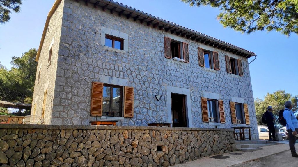 Muleta Gran-Refugio de la Muleta desde Sóller - Caminando por Mallorca Vista exterior del Refugio de la Muleta, una construcción de piedra con ventanas y persianas de madera, rodeado de vegetación.